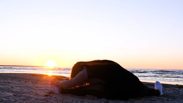 Молода жінка на тлі заходу сонця на березі моря займається вправами, бігаючи, розтягуючись і падаючи, Дівчина на морі робить йогу. Портрет: повільний рух, захід сонця
. - Фото, зображення