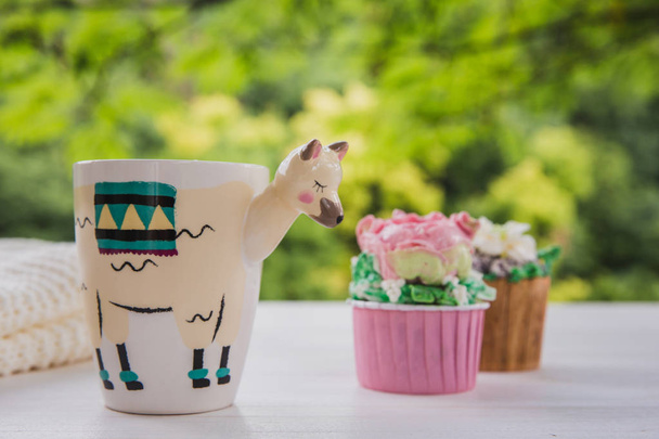 Beautiful llama shaped trendy cup with hot drink and two cupcakes on white wooden table with bright greens background - Foto, afbeelding