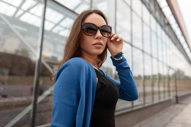 Melko houkutteleva nuori hipster nainen seksikäs huulet tyylikäs aurinkolasit tyylikäs sininen neulottu viitta muodikas musta pitsi alkuun kävelee lähellä moderni lasirakennus. Kaunis tyttö malli
. - Valokuva, kuva