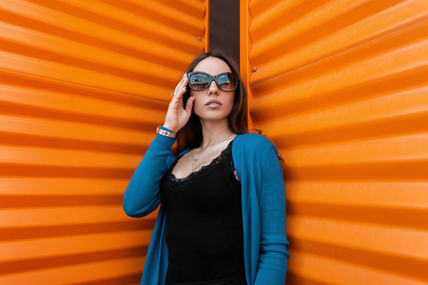 Beautiful young urban hipster woman in a vintage knitted blue cape in dark sunglasses in a fashionable black t-shirt is standing near the metallic orange wall outdoors. Cute girl model in the city. - Photo, Image
