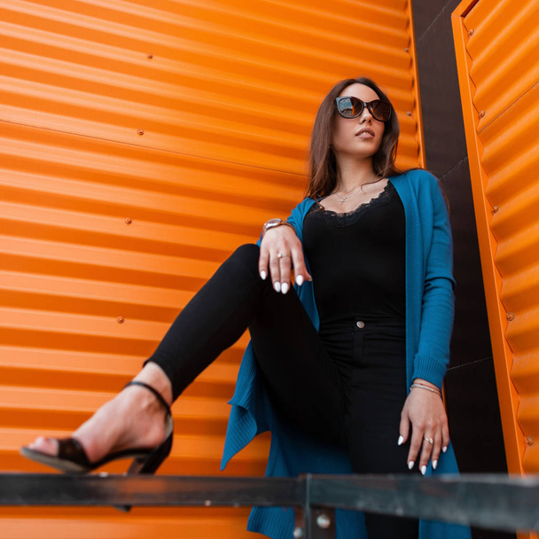 Pretty young hipster woman in stylish clothes in trendy sunglasses in trendy sandals poses near the modern orange metal wall in the city on a summer day. Attractive urban girl fashion model outdoors. - Photo, Image