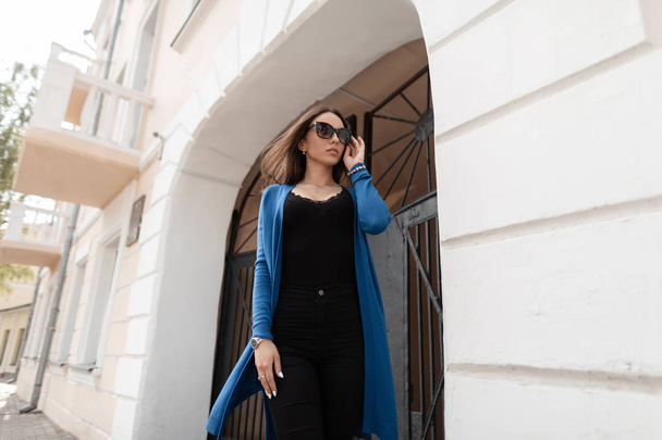 Die attraktive junge urbane Hipsterfrau in einem gestrickten blauen Umhang mit stylischer Sonnenbrille und modischem schwarzen T-Shirt steht neben einem alten Eisentor im Freien. schönes Mädchen Modell in der Stadt. Stil - Foto, Bild