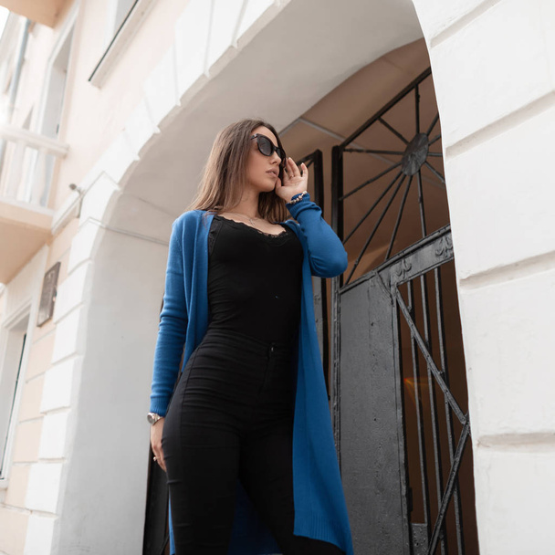 Stylish beautiful young hipster woman in sunglasses in a black T-shirt in a knitted summer cape in vintage jeans is standing near a vintage building with an iron gate in the city. Urban girl walks. - Foto, Bild