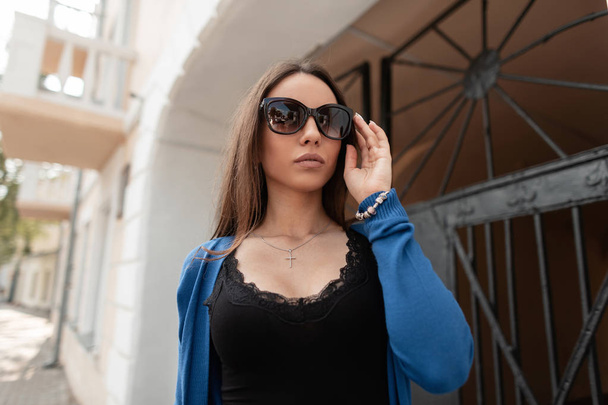 Pretty young urban hipster woman in a vintage knitted blue cape in dark sunglasses in a fashionable black t-shirt is standing near the vintage gate outdoors. Cute girl model walks in the city. - Foto, afbeelding