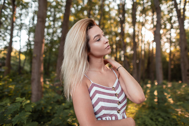 Göz alıcı pembe çizgili sundress güzel genç sarışın kadın güneşli sıcak bir yaz gününde ormanda ayakta rahatlatır. Çekici seksi fotoğraf modeli kız açık havada güneş sahiptir. Moda. - Fotoğraf, Görsel