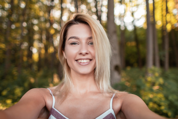 Young pretty joyful woman in a trendy pink dress makes selfie on a phone in the forest among the trees on a summer sunny day.Happy girl relaxes in nature and takes pictures of herself. - Photo, Image