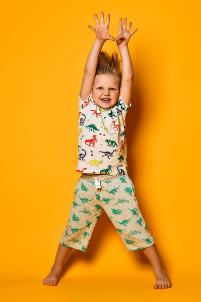 Little cute blond boy preschooler in pajamas with dinosaurs having fun has joy in the studio over yellow background. - Fotó, kép