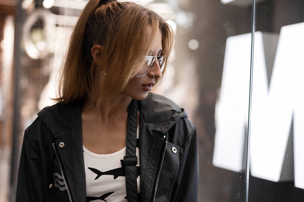Стильная рыжая молодая хипстерша с модной прической в модном пиджаке в футболке в винтажных очках возле яркой стеклянной стены в торговом центре. Европейская привлекательная модель
. - Фото, изображение