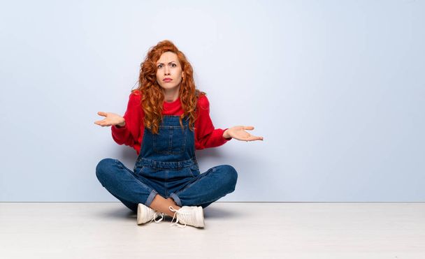 Femme rousse avec salopettes assis sur le sol malheureux pour ne pas comprendre quelque chose
 - Photo, image