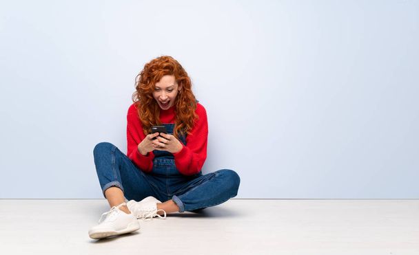 Redhead woman with overalls sitting on the floor surprised and sending a message - Фото, изображение