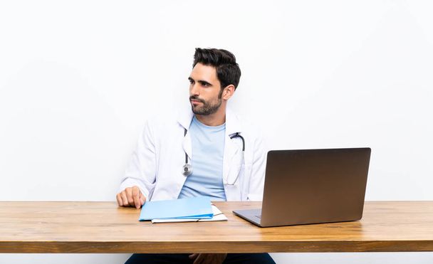 Jeune homme médecin avec son ordinateur portable sur mur isolé regardant côté
 - Photo, image