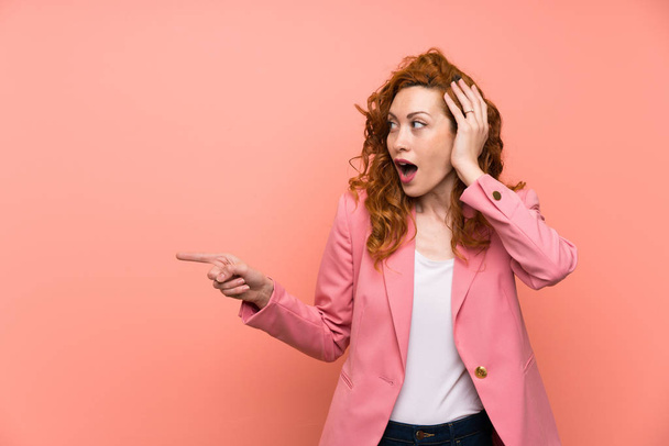 Redhead woman in suit over isolated pink wall surprised and pointing finger to the side - 写真・画像