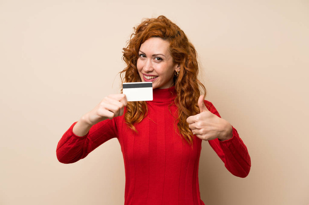 Redhead vrouw met coltrui trui houden van een creditcard - Foto, afbeelding