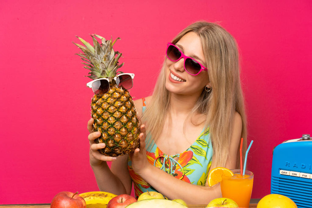 Young blonde woman in swimsuit holding a pineapple with sunglasses - Fotografie, Obrázek