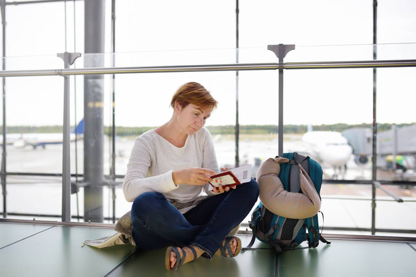 Reife Frau mit Rucksack und Reisekissen sitzt im Wartebereich des Flughafens und kontrolliert ihr Ticket. - Foto, Bild