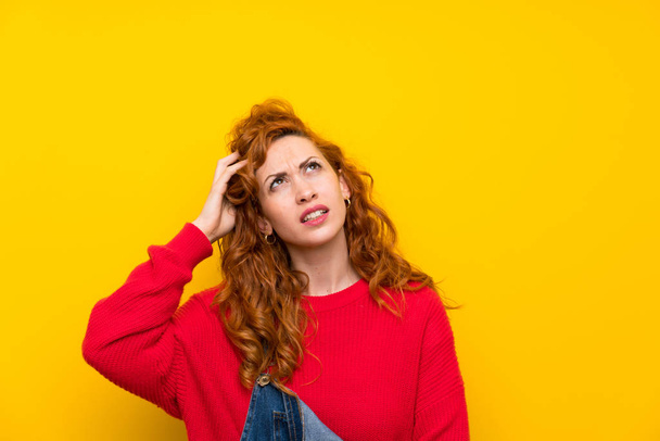 Femme rousse avec salopettes sur mur jaune isolé ayant des doutes et avec expression du visage confus
 - Photo, image