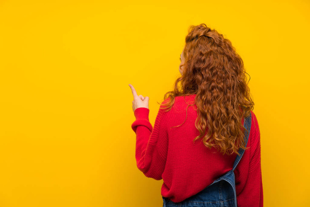 Redhead woman with overalls over isolated yellow wall pointing back with the index finger - Photo, Image