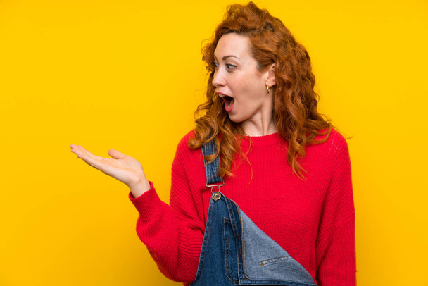 Redhead woman with overalls over isolated yellow wall holding copyspace imaginary on the palm - Photo, Image