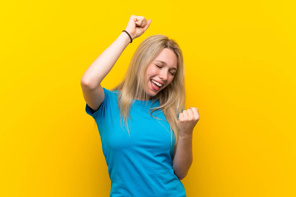 jonge blonde vrouw over geïsoleerde gele achtergrond vieren een overwinning - Foto, afbeelding