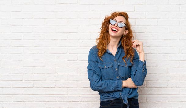 Redhead vrouw over witte bakstenen muur met bril en glimlachend - Foto, afbeelding