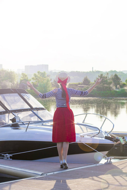 Bella ragazza in un cappello di paglia sul molo vicino al mare yacht e barche. Ragazza in gonna rossa e vestito da marinaio sul molo al tramonto in vacanza
 - Foto, immagini