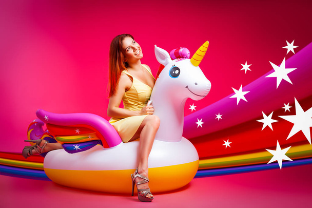 Concept summer mood, relaxation and beauty.Modern portrait of a young woman in a yellow dress resting on an inflatable unicorn mattress on an isolated pink background with rainbow and star - Photo, Image