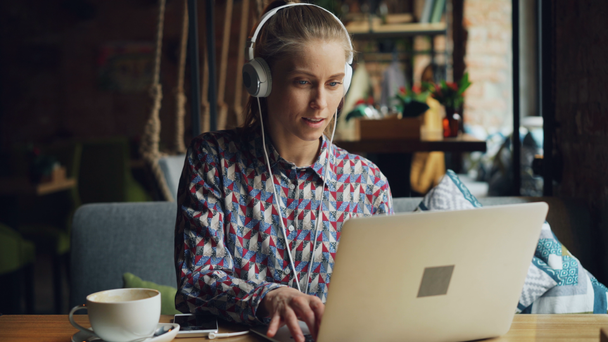 Χαρούμενο κορίτσι ακούγοντας μουσική στα ακουστικά χρησιμοποιώντας laptop πληκτρολόγηση τραγουδώντας στο καφέ - Πλάνα, βίντεο