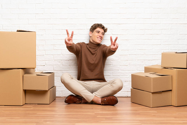 Knappe jonge man verhuizen in nieuw huis tussen dozen glimlachend en toont Victory teken - Foto, afbeelding