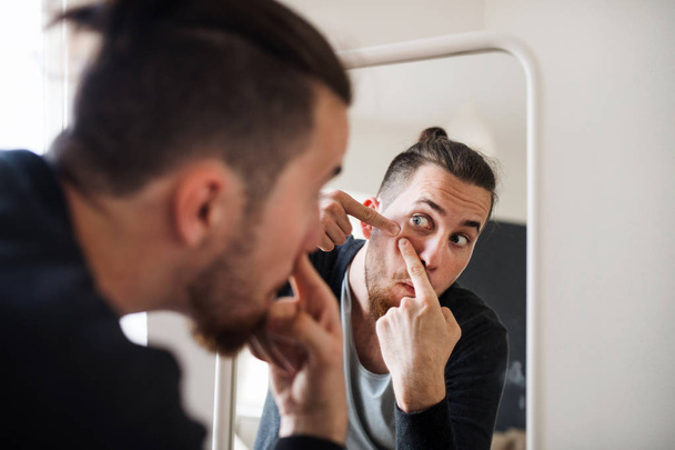 A young man indoors looking in the mirror, squeezing a pimple. - Foto, immagini