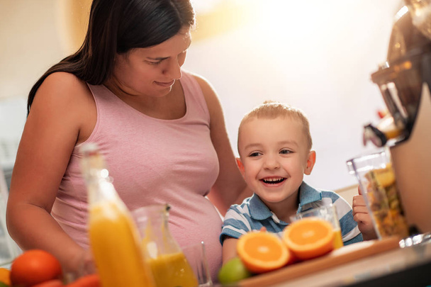 Zwangere moeder en haar zoon genieten in de keuken en maken vers sinaasappelsap. - Foto, afbeelding