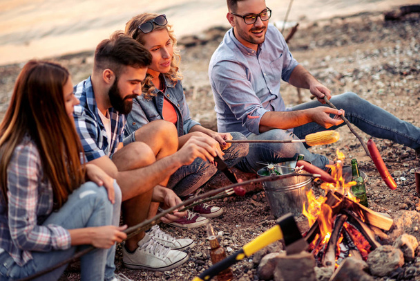 Gruppo di amici seduti intorno al fuoco al tramonto, grigliando salsicce e divertendosi sulla spiaggia. - Foto, immagini