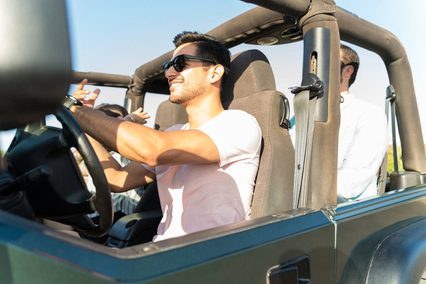 Happy young man with friends driving 4x4 vehicle during road trip on sunny day - Foto, imagen