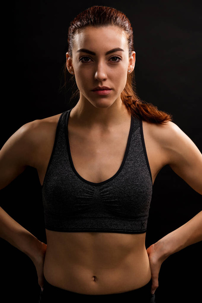 Portrait of confident young fit woman standing with hands on hip against black background - Φωτογραφία, εικόνα