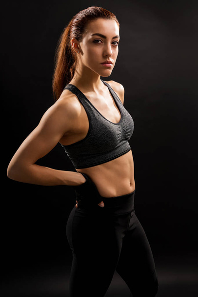 Portrait of pretty sportswoman standing with hands on hip against black background - Foto, afbeelding