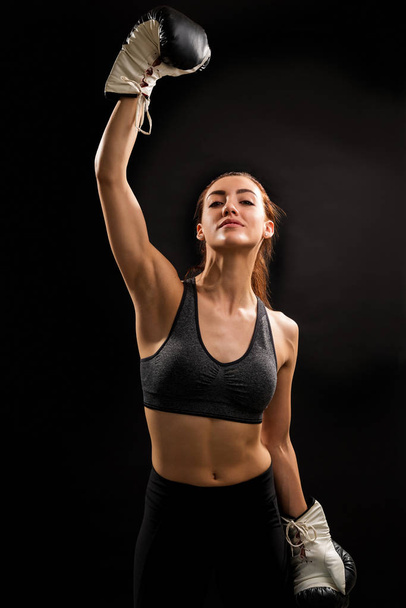 Confident young successful female boxer standing with arm raised against black background - Fotó, kép