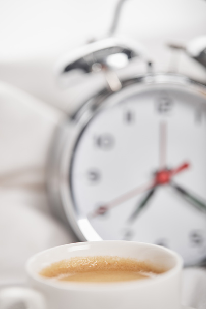 背景にベッドに銀の目覚まし時計と白いカップでコーヒーの選択的な焦点 - 写真・画像
