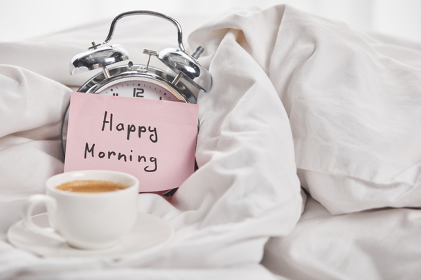 coffee in white cup near silver alarm clock with happy morning lettering on sticky note in bed - Foto, afbeelding
