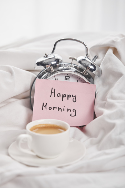 kávé fehér csésze csészealj közelében ezüst ébresztőóra boldog reggel betűkkel a ragadós cetlik az ágyban - Fotó, kép