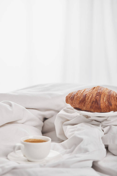 delicioso croissant na placa perto de café em copo branco no pires na cama
 - Foto, Imagem