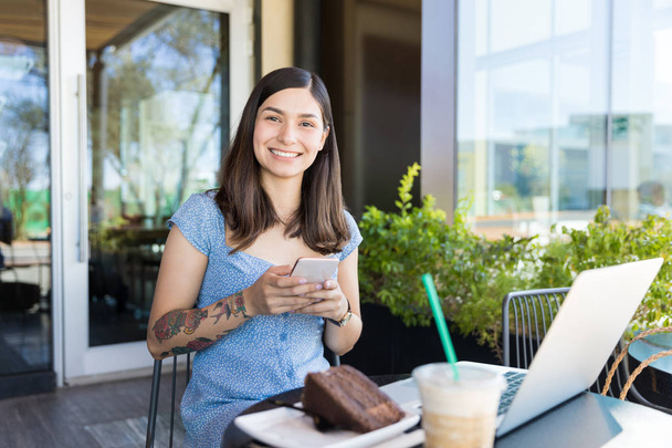 Portrait of confident female blogger using smartphone and laptop while having desert in cafe - Photo, Image