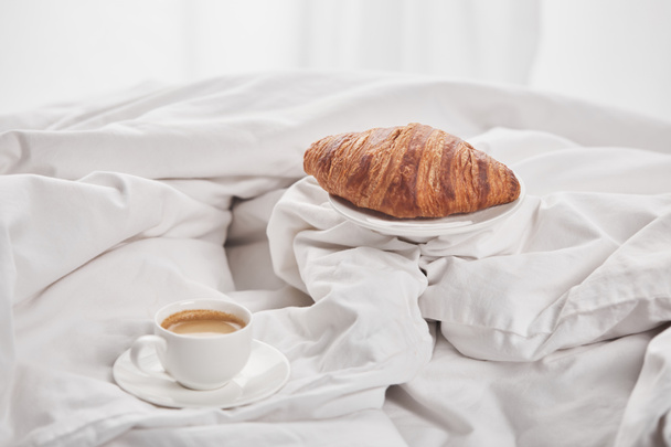 sabroso croissant en el plato cerca del café en la taza blanca en platillo en la cama
 - Foto, imagen