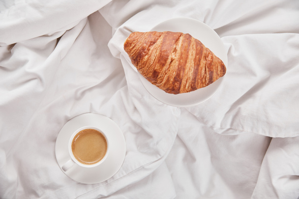 vista superior de croissant fresco en el plato cerca del café en taza blanca en platillo en la cama
 - Foto, Imagen