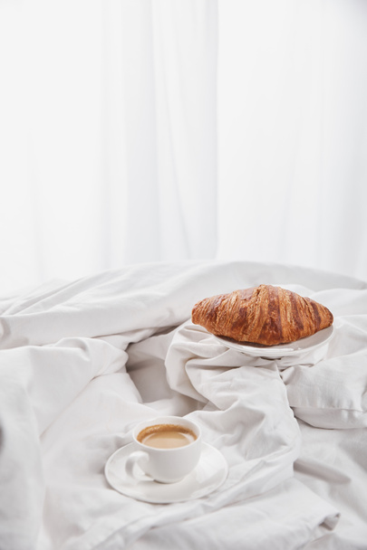 croissant fresco na placa perto do café na xícara branca no pires na cama
 - Foto, Imagem