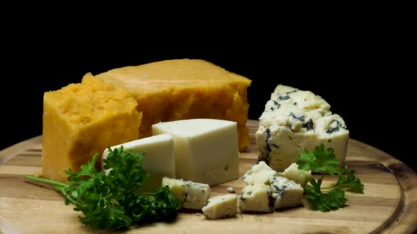 Close up for french delicious aged cheeses choped and served on wooden board isolated on black background. Frame. Cheddar, parmesan, and blue pieces of cheese with parsley. - Footage, Video