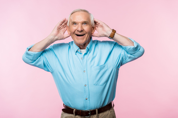 cheerful senior man touching ears and smiling while standing on pink  - Фото, зображення
