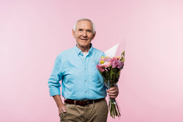 cheerful senior man standing with hand in pocket and holding bouquet on pink  - Photo, Image
