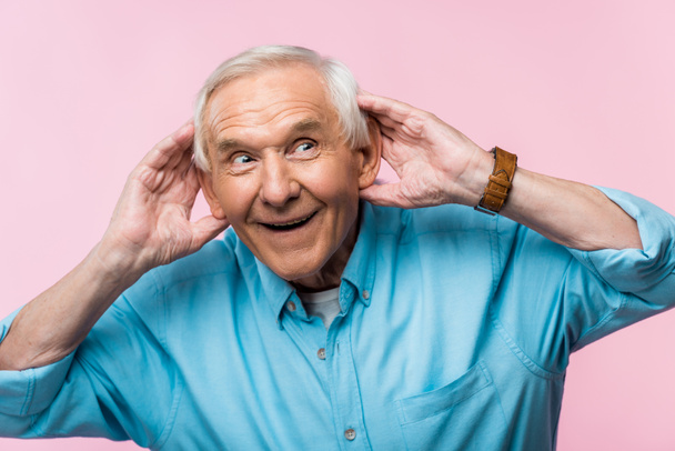 ευτυχισμένος ανώτερος άνθρωπος αγγίζοντας τα αυτιά και χαμογελά απομονωθεί σε ροζ  - Φωτογραφία, εικόνα