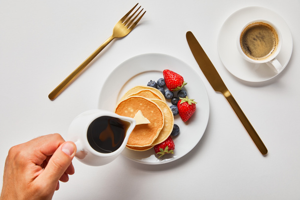 cropped view of man pouring syrup on pancakes with berries near golden cutlery and cup of coffee - Φωτογραφία, εικόνα