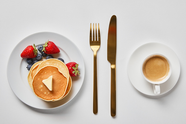 top view of tasty breakfast with pancakes, berries and cup of coffee near golden cutlery - Photo, Image