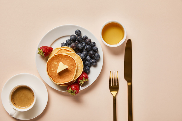 top view of breakfast with cup of coffee, pancakes, strawberries, blueberries and honey near golden cutlery - Fotografie, Obrázek